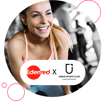Lächelnde Frau im Sporttop, die nach vorn lehnt, vor unscharfem Fitnessstudio‑Hintergrund; am unteren Bildrand die Logos „Edenred“ und „Urban Sports Club“.