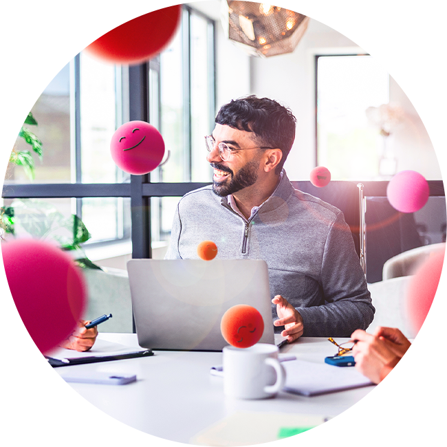Person sitzt in einem modernen Büro an einem Laptop, umgeben von schwebenden pinken Kugeln mit lächelnden Symbolen. Auf dem Tisch liegen Unterlagen, Stifte und eine Tasse, während im Hintergrund große Fenster für helles Tageslicht sorgen