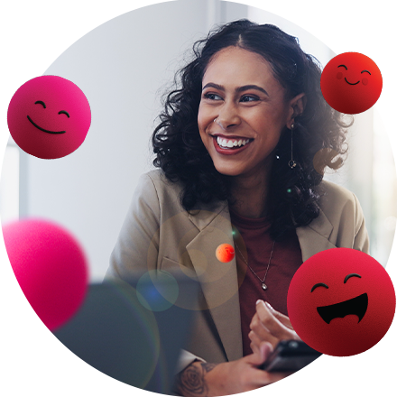 Frau sitzt lächelnd in einem hellen Raum und hält ein Smartphone in der Hand. Um sie herum schweben mehrere pinke Kugeln mit fröhlichen Emoji‑Gesichtern.