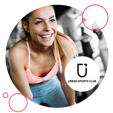 Lächelnde Frau im Sporttop, die nach vorn lehnt, vor unscharfem Fitnessstudio‑Hintergrund; am unteren Bildrand ist das Logo  „Urban Sports Club“ zu sehen.